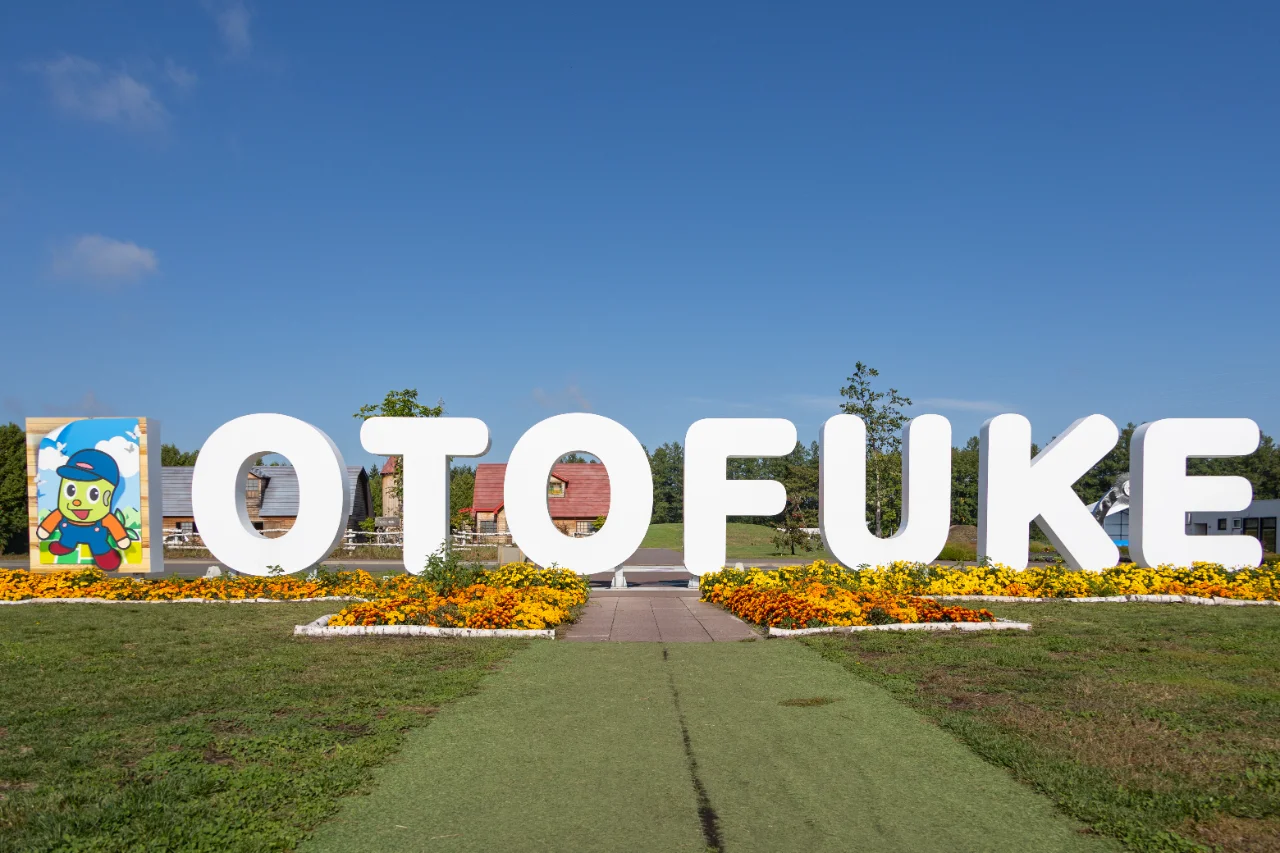 青空の下で花に囲まれたOTOFUKEのモニュメントと背景のなつぞらエリアの建物